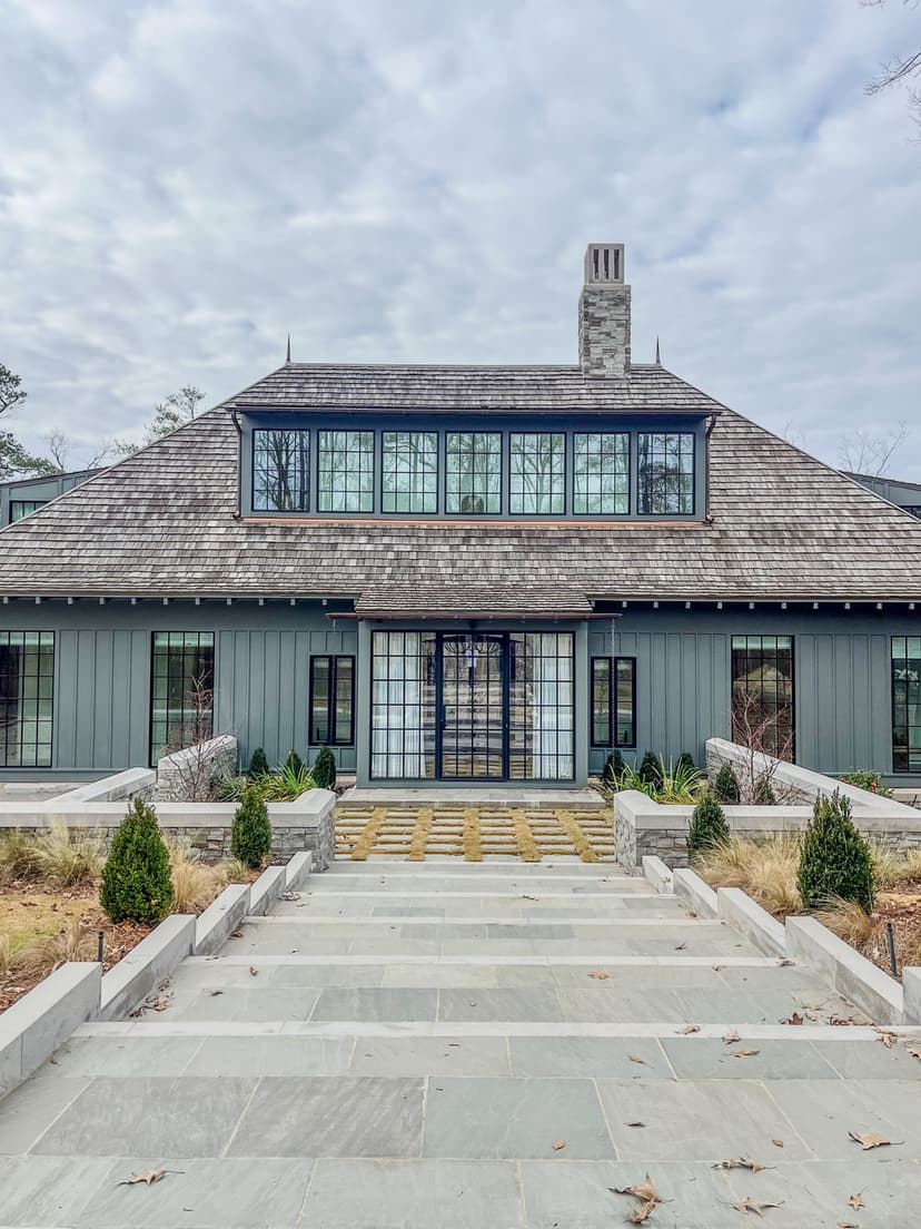 Modern gray house with large windows and stone walkway, surrounded by manicured landscaping.